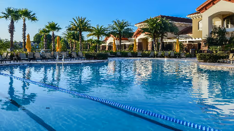 Outdoor swimming pool with lounge chairs, umbrellas, and palm trees in front of a Mediterranean-style building.