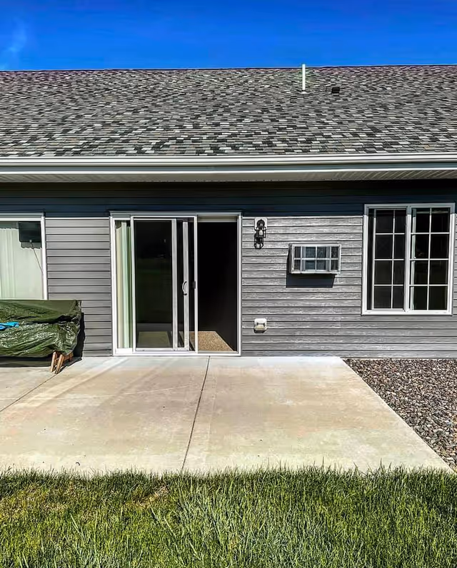 Gray single-story building exterior with sliding glass door open to a concrete patio and blue sky.