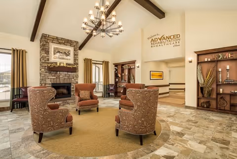 A spacious and well-lit living room area with four patterned armchairs arranged in a circle on a round rug. The room features a stone fireplace with a framed picture above it, large windows with curtains, wooden beams on the ceiling, a modern chandelier, and built-in wooden shelves with decorative items. A sign on the wall reads 'Welcome to Advanced Rehabilitation & Healthcare of Bowie.'