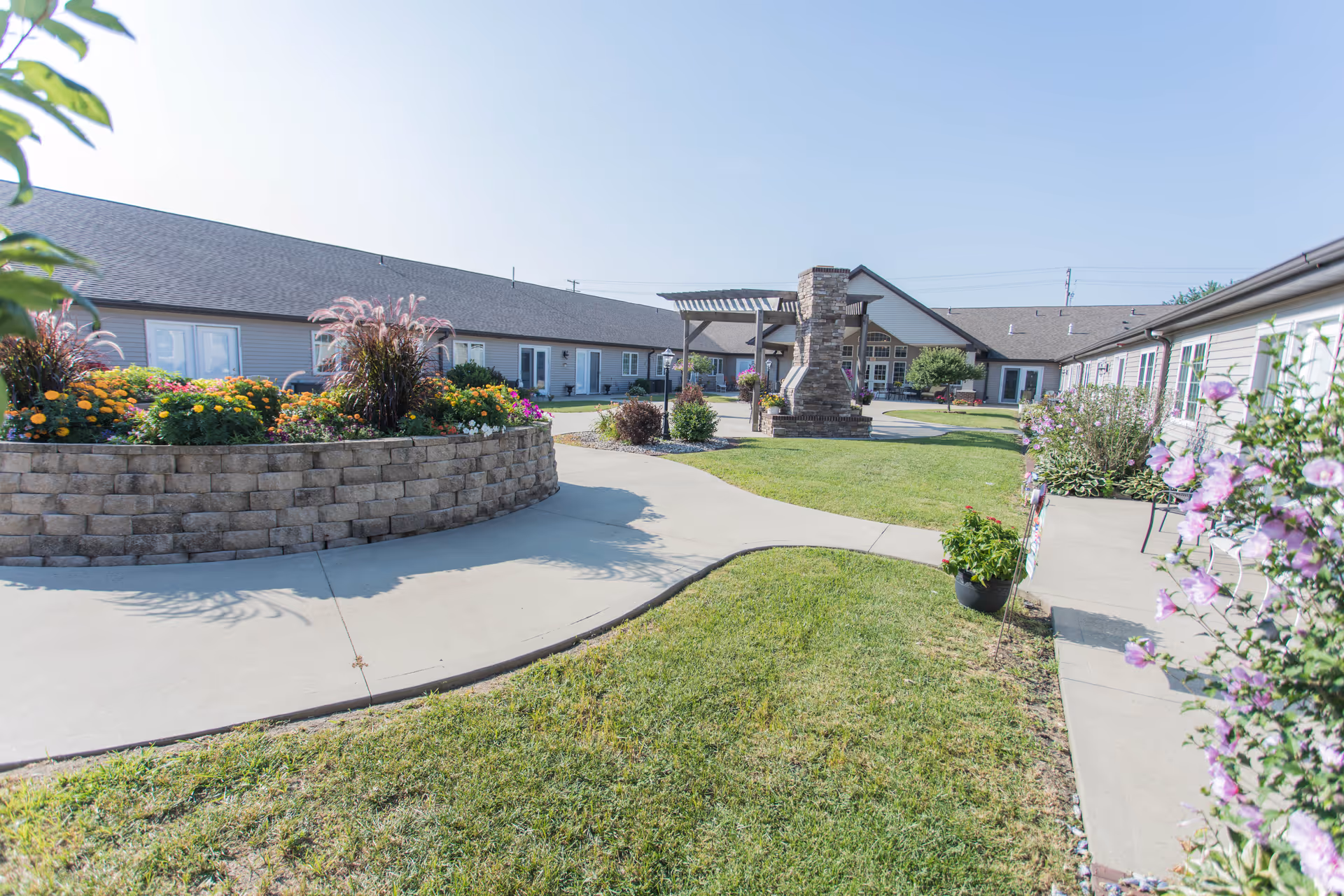 Sunny courtyard with raised flowerbeds, paved walkways, lawn, and a pergola between single-story buildings.