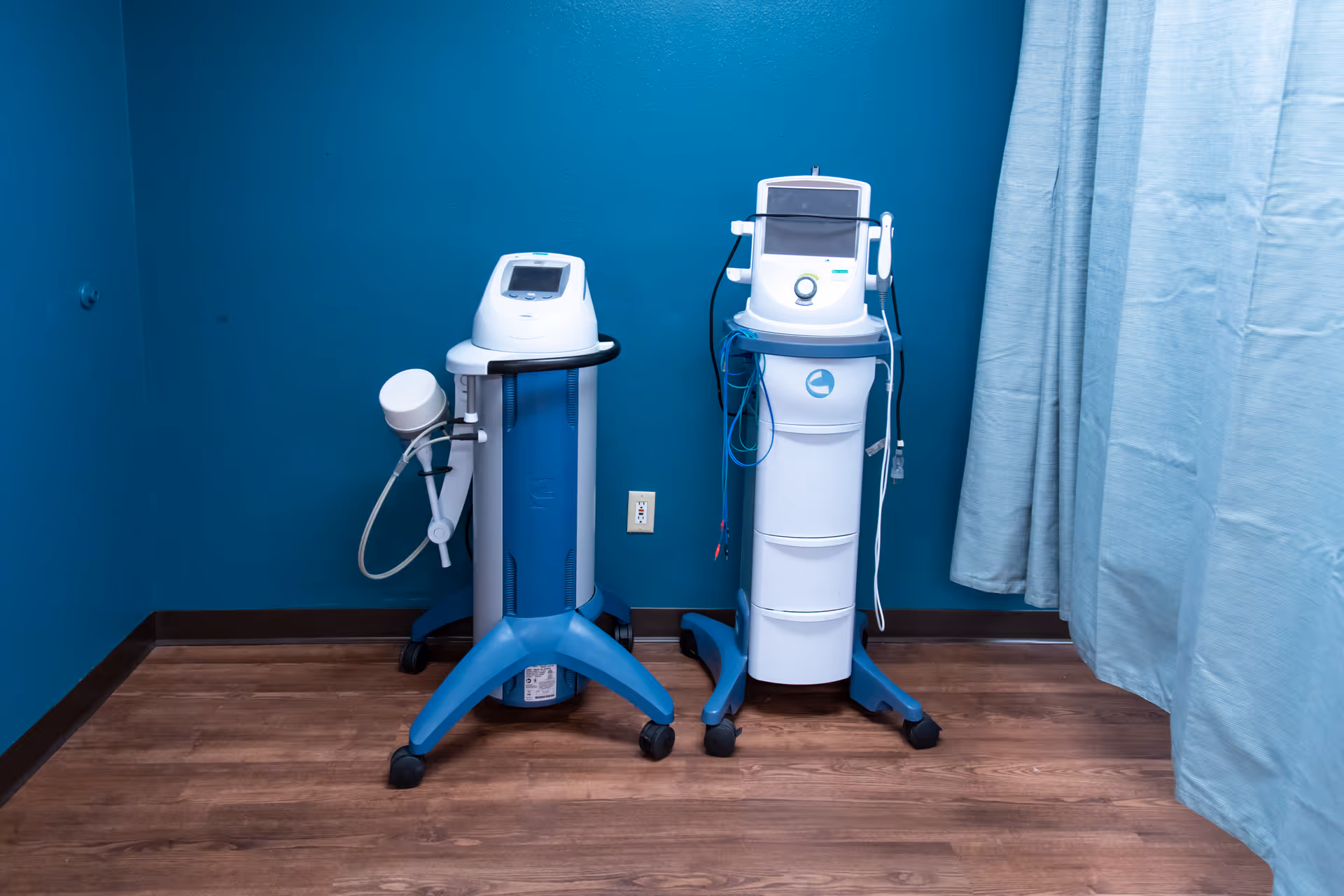Two wheeled medical devices standing against a teal wall with wood flooring and a light blue curtain to the right.
