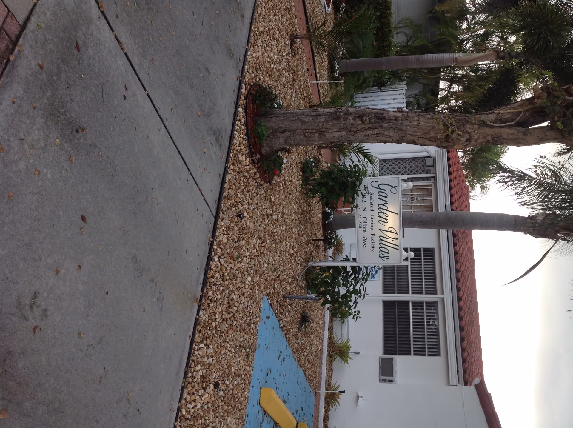 Exterior front of a single-story white building with palm trees, gravel landscaping, a blue handicap parking marker, and a sign reading 'Garden Villas'.