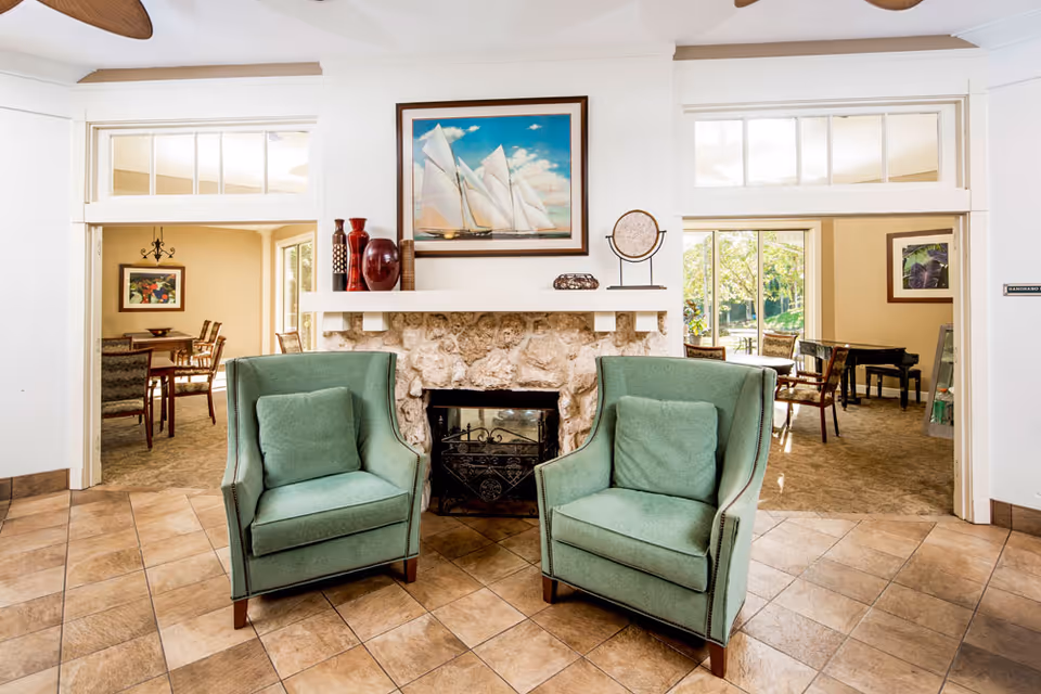 A cozy interior space featuring two green armchairs placed in front of a stone fireplace with a framed painting of sailboats above it. On the mantel are decorative vases and a small sculpture. To the left is a dining area with a table and chairs, and to the right is a room with a piano and additional seating, with large windows letting in natural light.