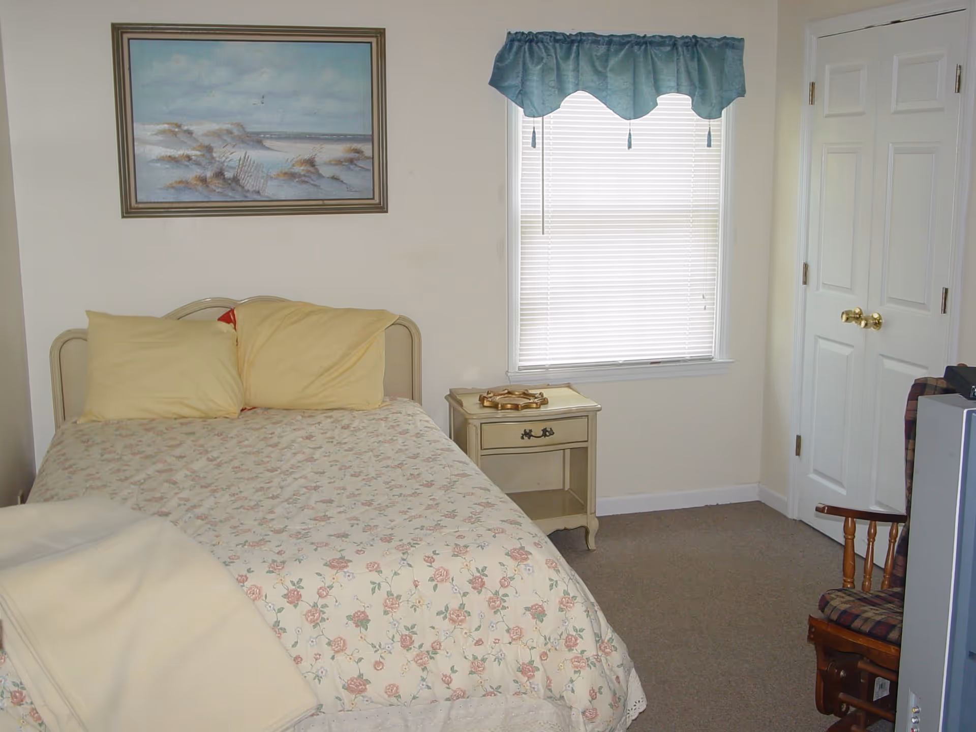 A small bedroom with a bed covered in floral bedding and two yellow pillows. There is a nightstand with a decorative item next to the bed, a window with closed blinds and a blue valance, a framed painting of a beach scene on the wall, a closed white door with gold knobs, and a wooden rocking chair with a plaid cushion partially visible next to a television.