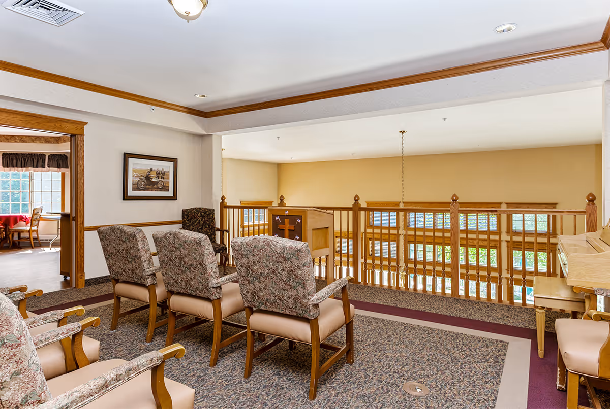 A small seating area with floral upholstered chairs arranged in rows facing a wooden podium with a cross on it, located on an upper level overlooking a lower floor with large windows. The space has carpeted floors, wood trim, and a piano on the right side. An adjacent room with a table and chairs is visible through an open doorway.