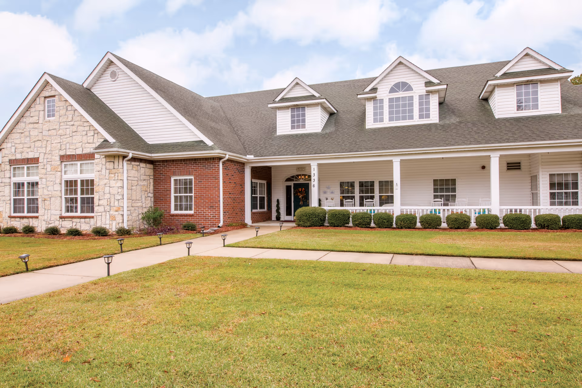 Front exterior view of a senior living facility building with a combination of stone, brick, and white siding. The building has a covered porch with white railings, multiple windows, and dormer windows on the roof. There is a well-maintained lawn and a concrete walkway leading to the entrance.