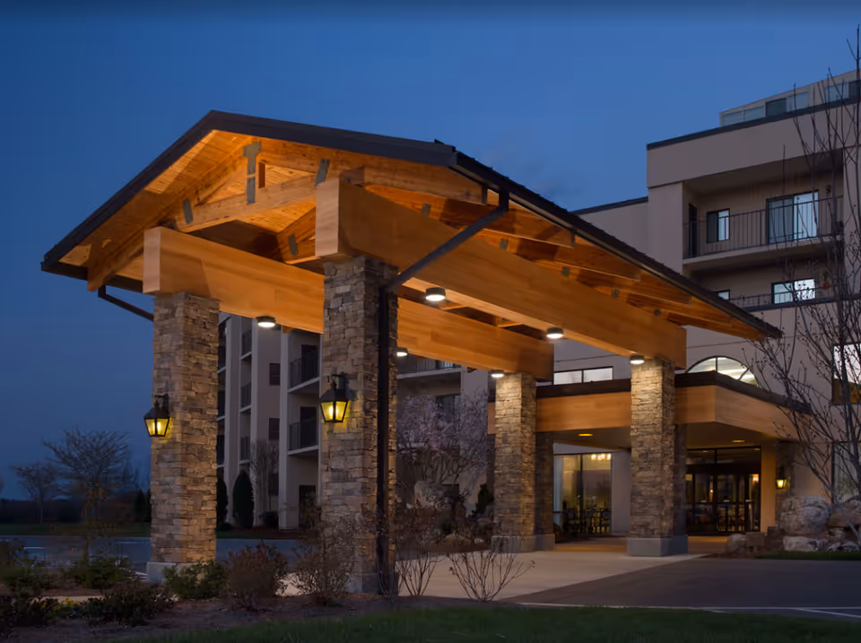 Entrance of Grace Ridge Retirement Community at dusk featuring a covered driveway with wooden beams and stone pillars illuminated by warm lights, with the building visible in the background.