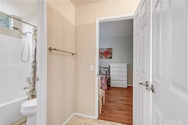 View of a small hallway area with a bathroom on the left featuring a bathtub, shower, toilet, and a floral shower curtain. The hallway leads to a bedroom with wooden flooring, a white dresser, a bed with a floral bedspread, and a floral wall decoration.