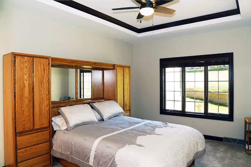 A bedroom with a large bed covered in gray and white bedding, two pillows, a wooden headboard with built-in cabinets and a mirror, a window with black trim letting in natural light, and a ceiling fan with a light fixture.