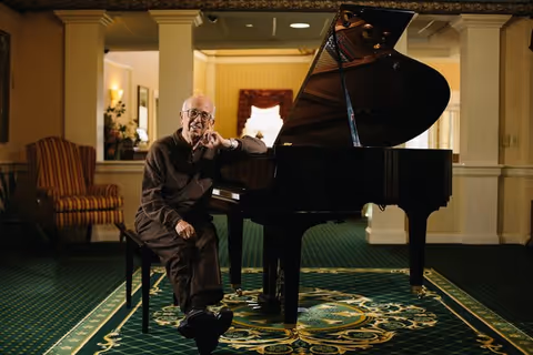 An elderly man sitting on a bench next to a grand piano in a warmly lit room with green carpet and patterned rug. The room has cream-colored walls, a striped armchair, and a window with a red curtain in the background.