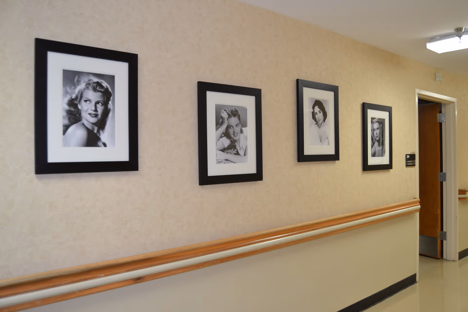 A hallway in a senior living facility with beige walls and a wooden handrail. Four black-framed black-and-white portraits of women are hung on the wall. There is an open door at the end of the hallway.
