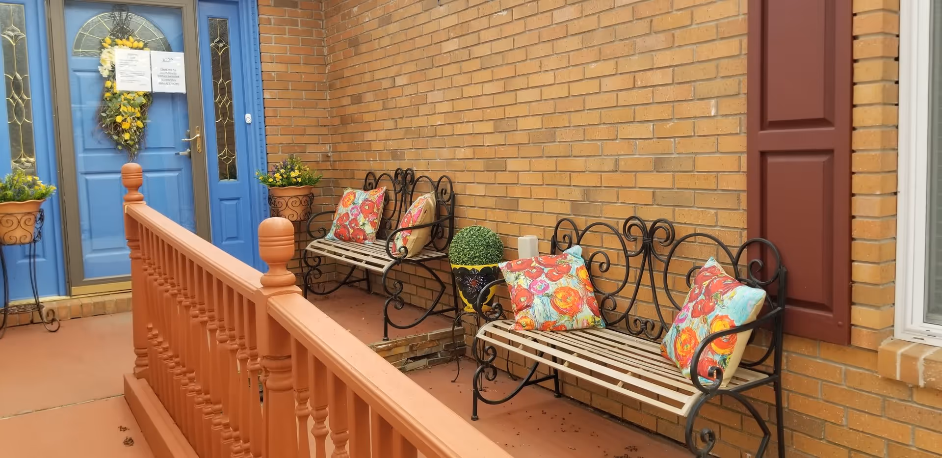 Outdoor seating area with two metal benches adorned with colorful floral cushions, a brick wall, a blue door decorated with a yellow flower wreath, and potted plants on either side of the door.