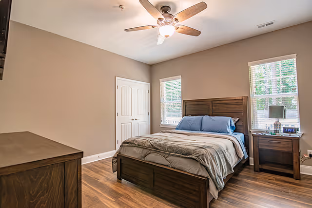 Well-lit bedroom with a wooden bed, nightstand and dresser, hardwood floors, ceiling fan, and two windows.