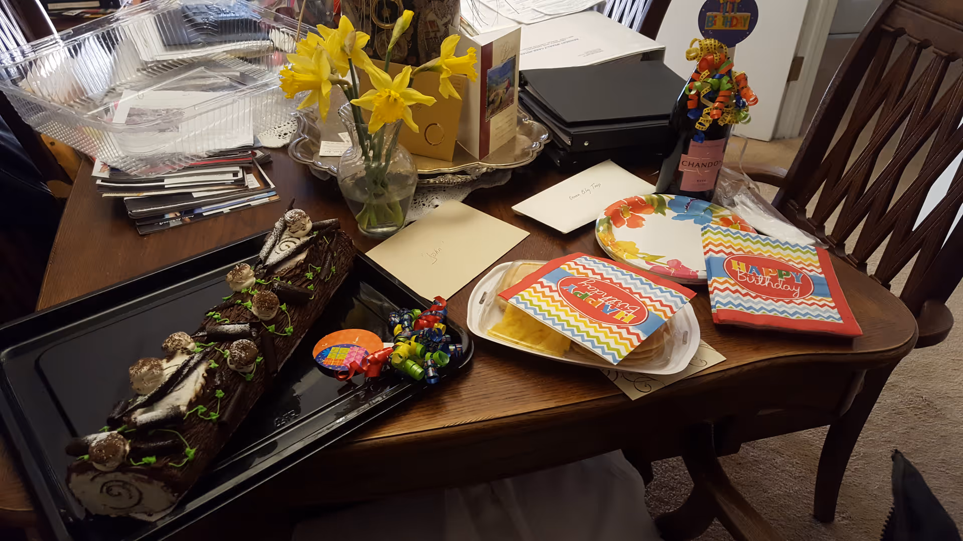 Wood dining table with a chocolate log cake, vase of daffodils, party plates, 'Happy Birthday' napkins and greeting cards.