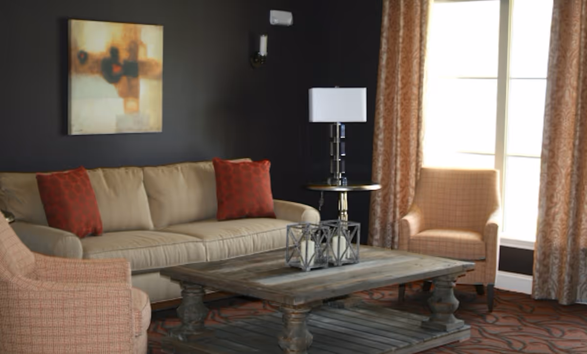 A cozy living room with a beige sofa adorned with two red patterned pillows, two upholstered armchairs, a rustic wooden coffee table with decorative candle holders, a side table with a modern lamp, dark walls, patterned curtains, and a large window letting in natural light.