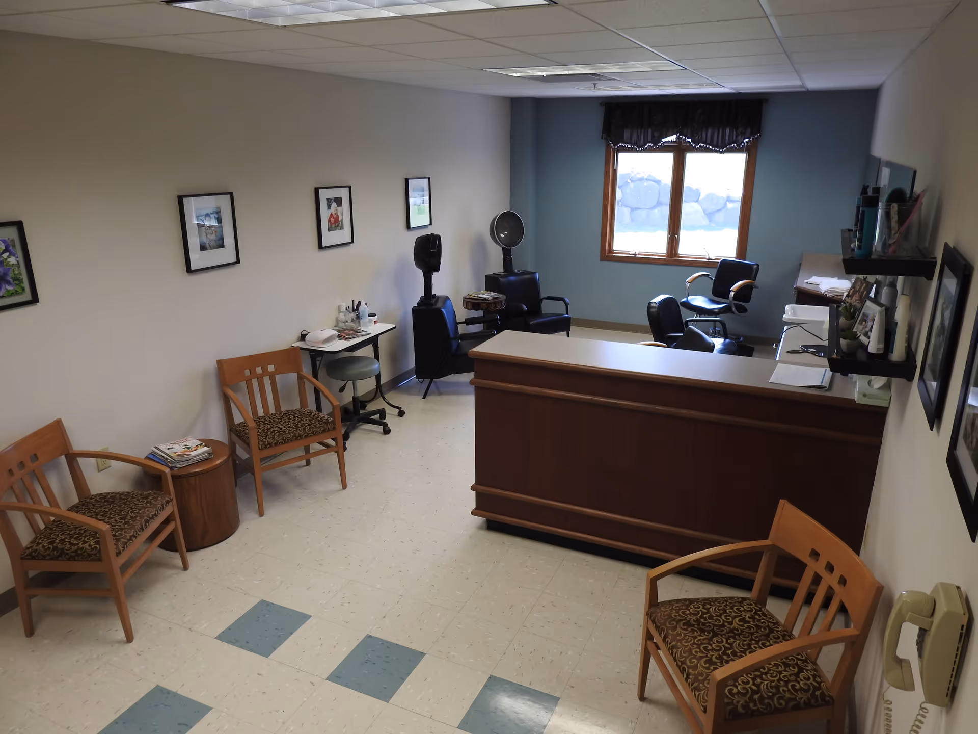 Interior view of a salon area in Coventry Village with several chairs, a reception desk, hair drying stations, framed pictures on the walls, and a window with a dark valance letting in natural light.