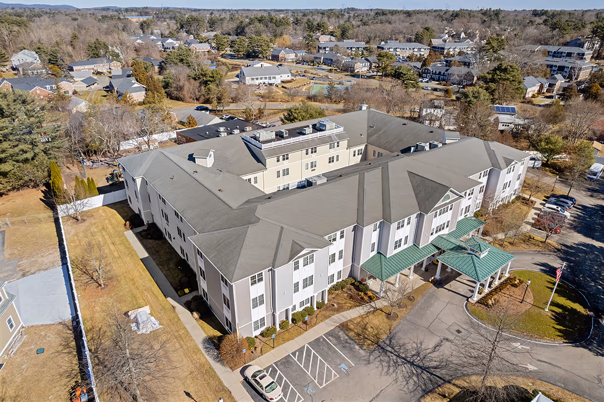 Aerial view of a large senior living facility building with a green roof entrance canopy, surrounded by parking spaces and landscaped grounds. The building is situated in a suburban area with other houses and trees visible in the background.