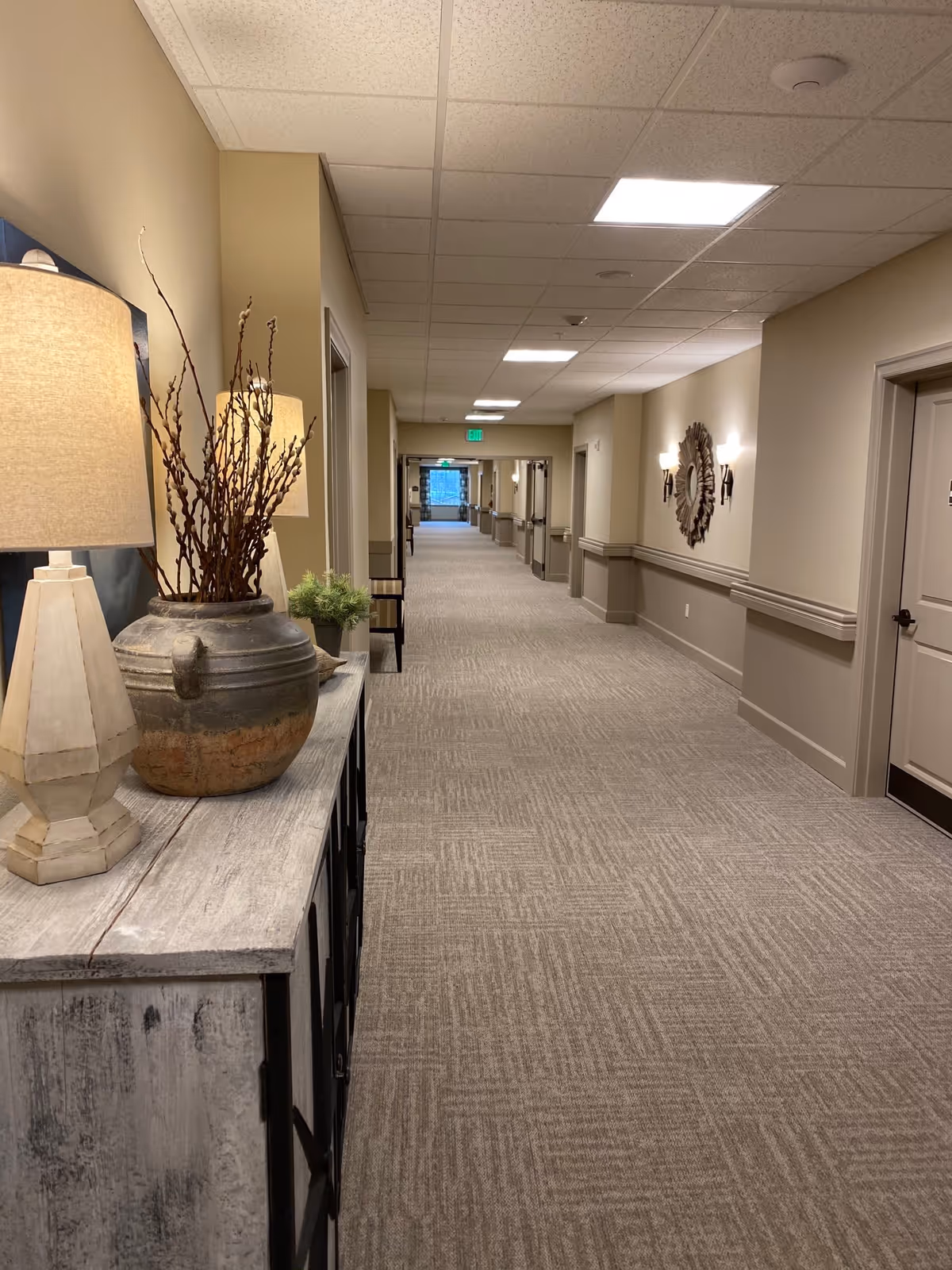 A long, well-lit hallway in a senior living facility with beige walls and carpeted floor. On the left side, there is a rustic wooden console table with a lamp, a large ceramic vase with decorative branches, and a small plant. The hallway has several doors on both sides and a decorative mirror with wall sconces on the right wall. The ceiling has recessed lighting panels.
