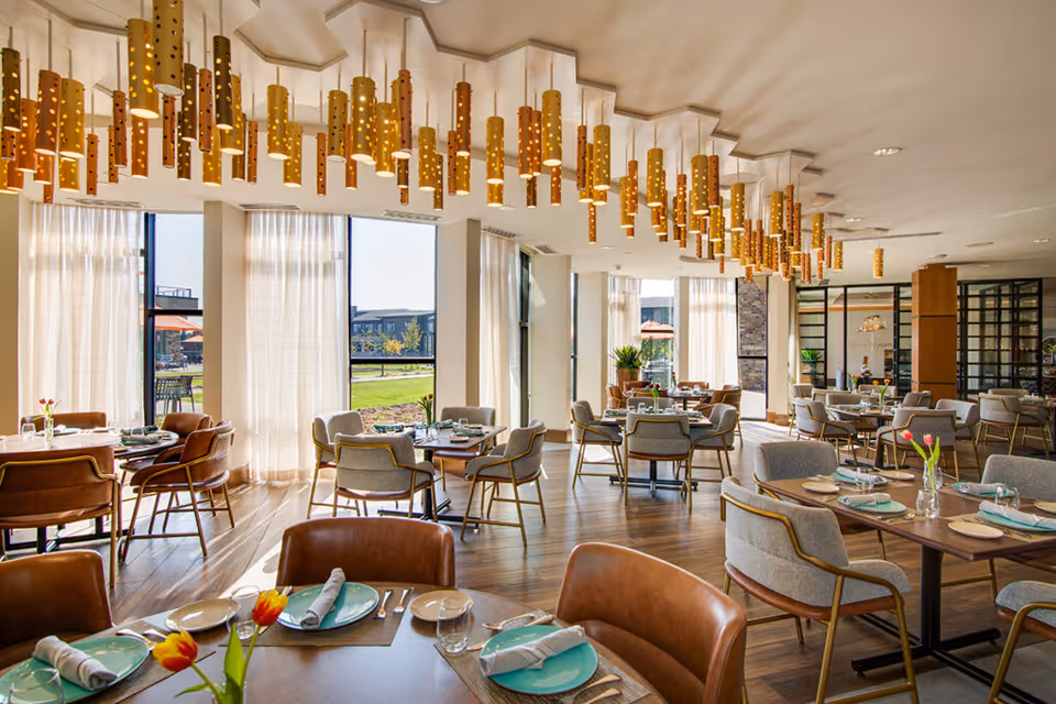 A bright and spacious dining room with large windows letting in natural light. The room features multiple tables set with plates, napkins, glasses, and cutlery. Chairs with leather and fabric upholstery surround the tables. Modern cylindrical pendant lights hang from the ceiling, and there are vases with tulips on some tables. Outside the windows, a green lawn and buildings are visible.