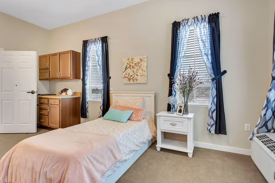 A cozy senior living bedroom with a single bed covered in floral bedding and a pink blanket. The room has two windows with blue and white curtains, a white nightstand with a vase of flowers and a framed photo, and wooden cabinets with a countertop on one side. The walls are light beige, and there is a white door partially open.