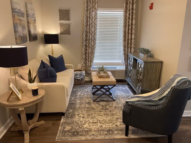 A cozy living room with a beige sofa adorned with dark blue pillows, a wooden side table with a lamp and small plant, a patterned area rug, a cushioned ottoman coffee table, a dark upholstered armchair with a striped throw, a wooden cabinet with decorative items, and a window with patterned curtains.