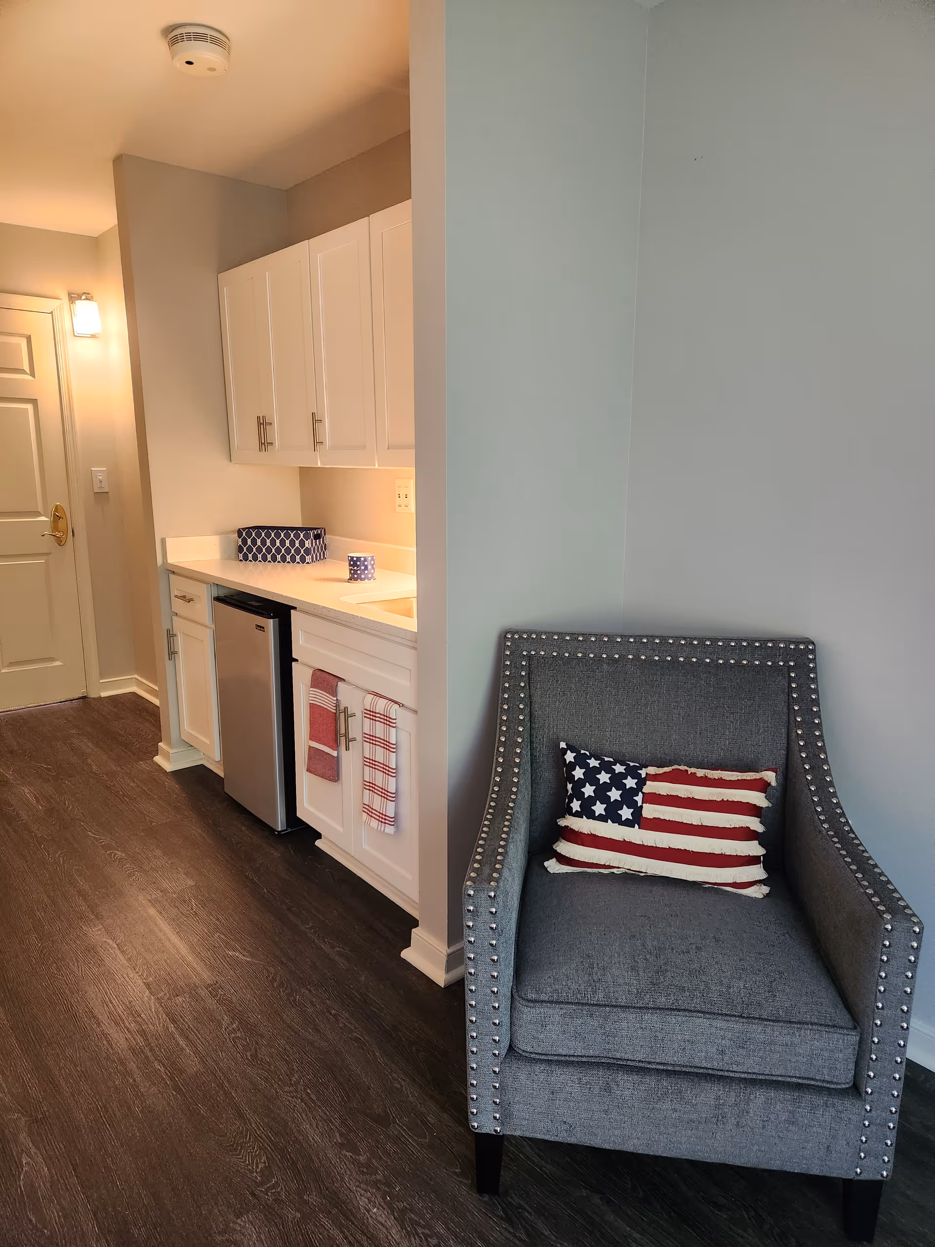A small kitchenette area with white cabinets, a mini refrigerator, and a countertop with a blue patterned container and a mug. Next to the kitchenette is a gray upholstered armchair with silver studs along the edges, featuring a decorative pillow with an American flag design. The floor is dark wood, and there is a closed door with a gold handle in the background.