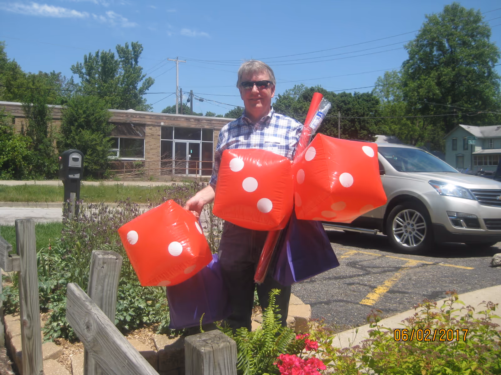 A man wearing sunglasses and a checkered shirt is standing outdoors on a sunny day, holding three large inflatable red dice and some purple bags. Behind him is a parking lot with a silver SUV and a building with large windows. There are plants and flowers in the foreground.