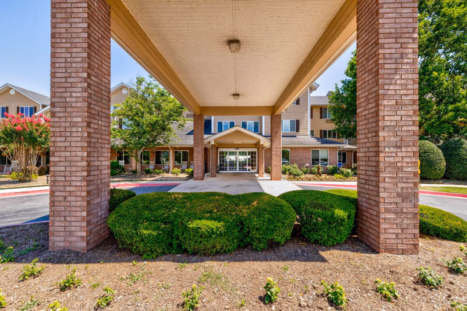 Covered entrance with brick columns and trimmed shrubs leading to the front doors of a multi-story senior living building.