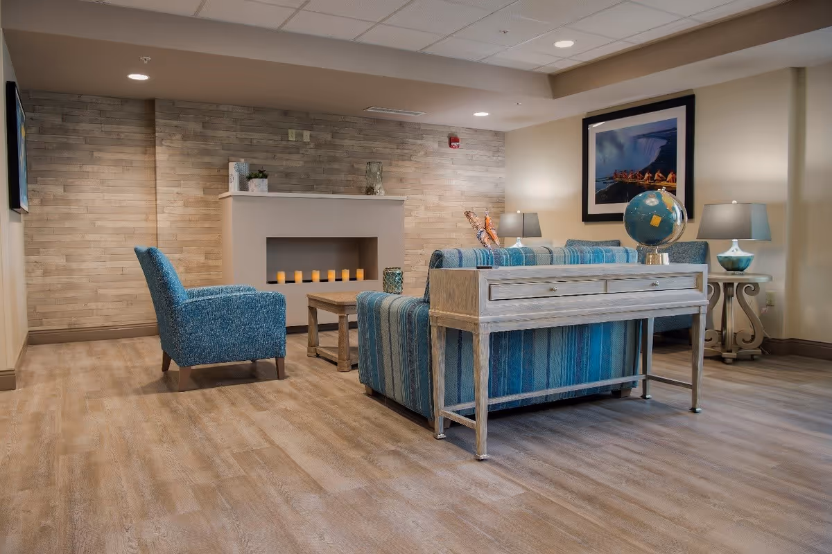 A cozy living room area with a blue striped sofa, a matching blue armchair, a wooden coffee table, and a console table with a globe on it. The room features a modern fireplace with candles inside, two table lamps on side tables, wood-style flooring, and a framed picture on the wall.