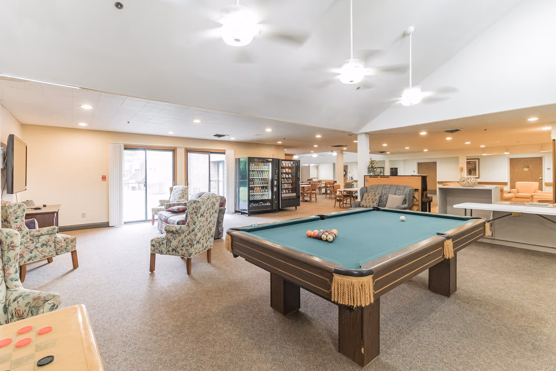 A spacious common area in a senior living facility featuring a pool table with balls arranged for a game, several floral upholstered armchairs, a vending machine, a television mounted on the wall, and multiple tables and chairs in the background. The room is well-lit with ceiling lights and ceiling fans, and has large sliding glass doors letting in natural light.
