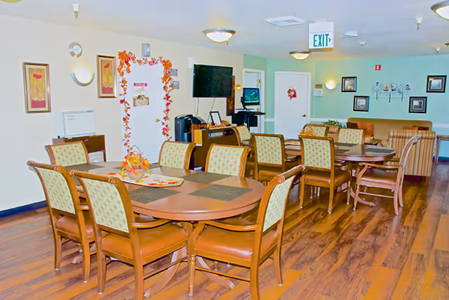 A communal dining area with multiple wooden tables and cushioned chairs arranged neatly. The room has hardwood flooring, light-colored walls adorned with framed artwork, and ceiling lights. There is a television mounted on the wall and an exit sign above a door in the background. Some autumn-themed decorations are visible on a door.