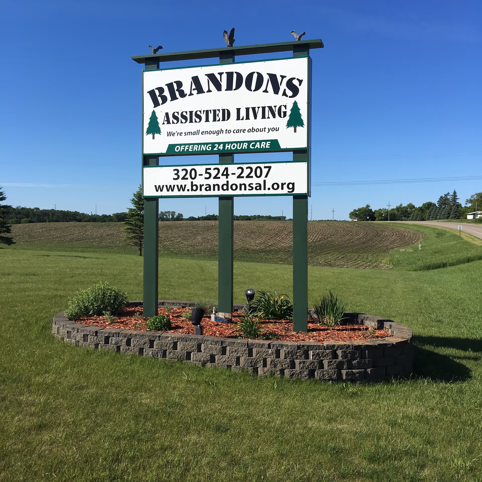 A large outdoor sign for Brandons Assisted Living standing on a landscaped area with green grass and small plants. The sign includes the text 'BRANDONS ASSISTED LIVING We're small enough to care about you OFFERING 24 HOUR CARE' along with a phone number and website URL. The background shows a clear blue sky and a rural landscape with fields and trees.