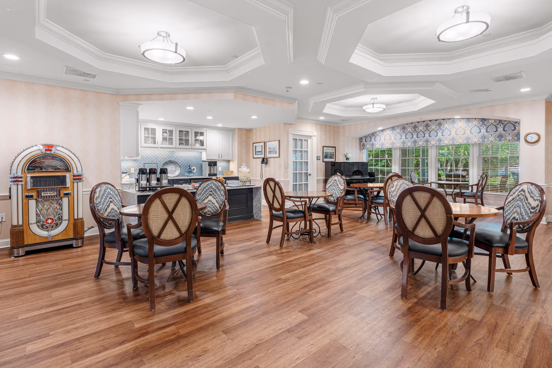 A spacious dining room with wooden floors, multiple round tables with chairs, a vintage jukebox on the left, a small kitchen area with coffee dispensers, and large windows with patterned valances letting in natural light. There is a piano near the windows and framed pictures on the walls.