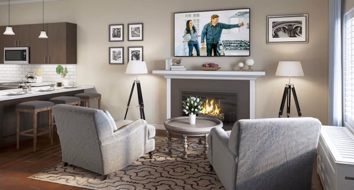 A cozy living room with two gray armchairs facing a round wooden coffee table with a vase of white flowers. Behind the table is a fireplace with a fire burning and a flat-screen TV mounted above it showing a man and woman in a kitchen. The room has two tripod floor lamps with white shades, framed black and white artwork on the walls, and a large window with blinds letting in natural light. To the left is an open kitchen with dark wood cabinets, a microwave, and a breakfast bar with three stools.
