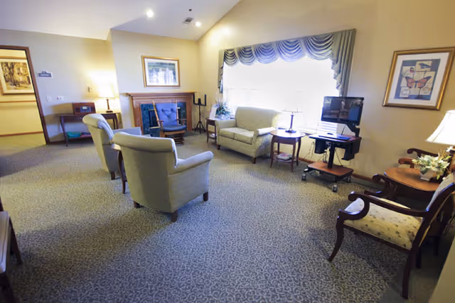 A cozy living room area with several upholstered chairs and a loveseat arranged around a small wooden table and a fireplace. The room has a large window with draped curtains letting in natural light, a TV on a rolling stand, framed artwork on the walls, and soft carpeted flooring.