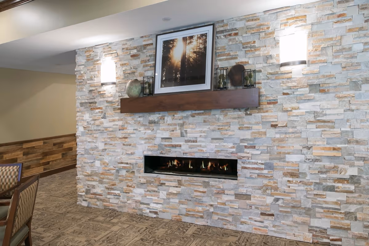 Interior view of a cozy room featuring a stone accent wall with a modern rectangular fireplace built into it. Above the fireplace is a wooden mantel holding decorative items and a framed picture of sunlight filtering through trees. Two wall-mounted lights illuminate the stone wall. Part of a dining table and chairs with patterned upholstery are visible on the left side.