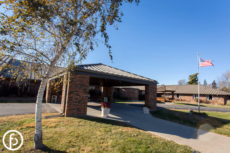 Brick single-story health and rehabilitation facility entrance with a covered drop-off, American flag, and a birch tree in front.
