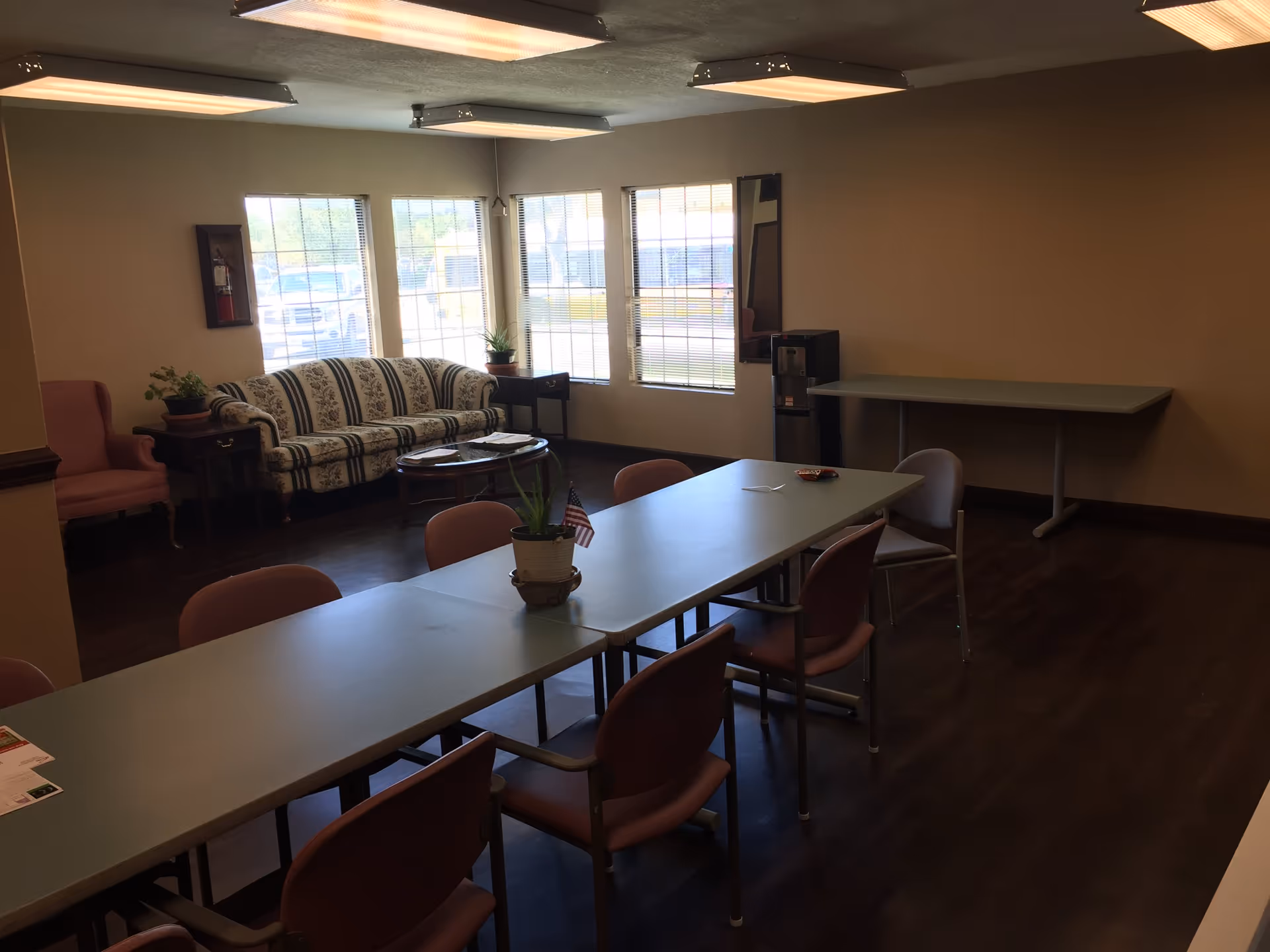 A common room with a long table surrounded by chairs, a small potted plant with an American flag on the table, a floral patterned sofa, a pink armchair, two small side tables with plants, large windows letting in natural light, a water dispenser, and a folded table against the wall.