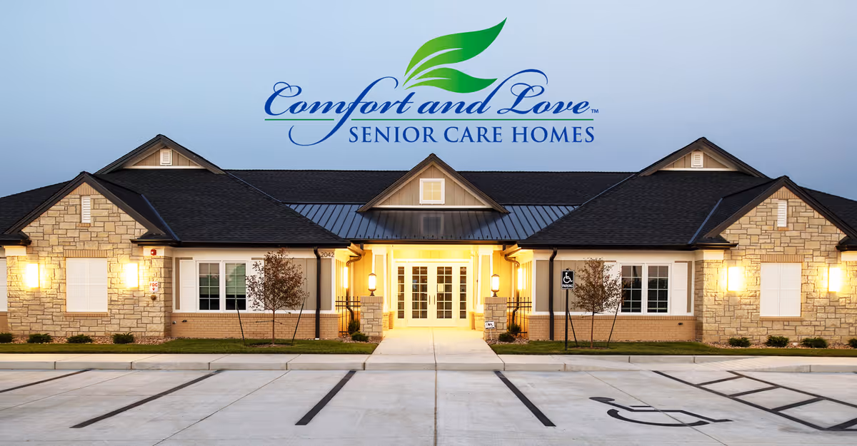 Front exterior of a single-story senior care facility with a lit central entrance, parking spaces, and a large 'Comfort and Love Senior Care Homes' logo above.