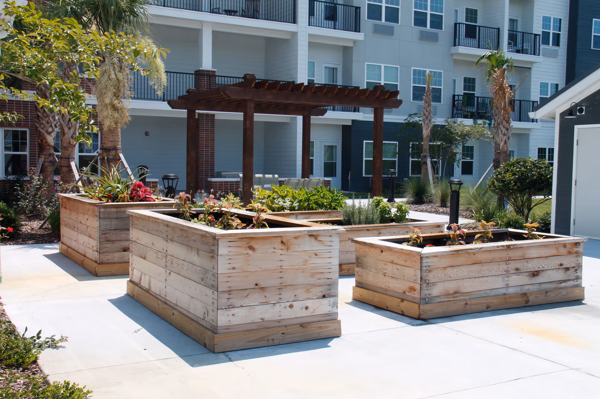 Outdoor garden area with several raised wooden planter boxes containing various plants and flowers. In the background, there is a wooden pergola with seating underneath, surrounded by palm trees and other greenery. The setting is part of a multi-story residential building with balconies and large windows.