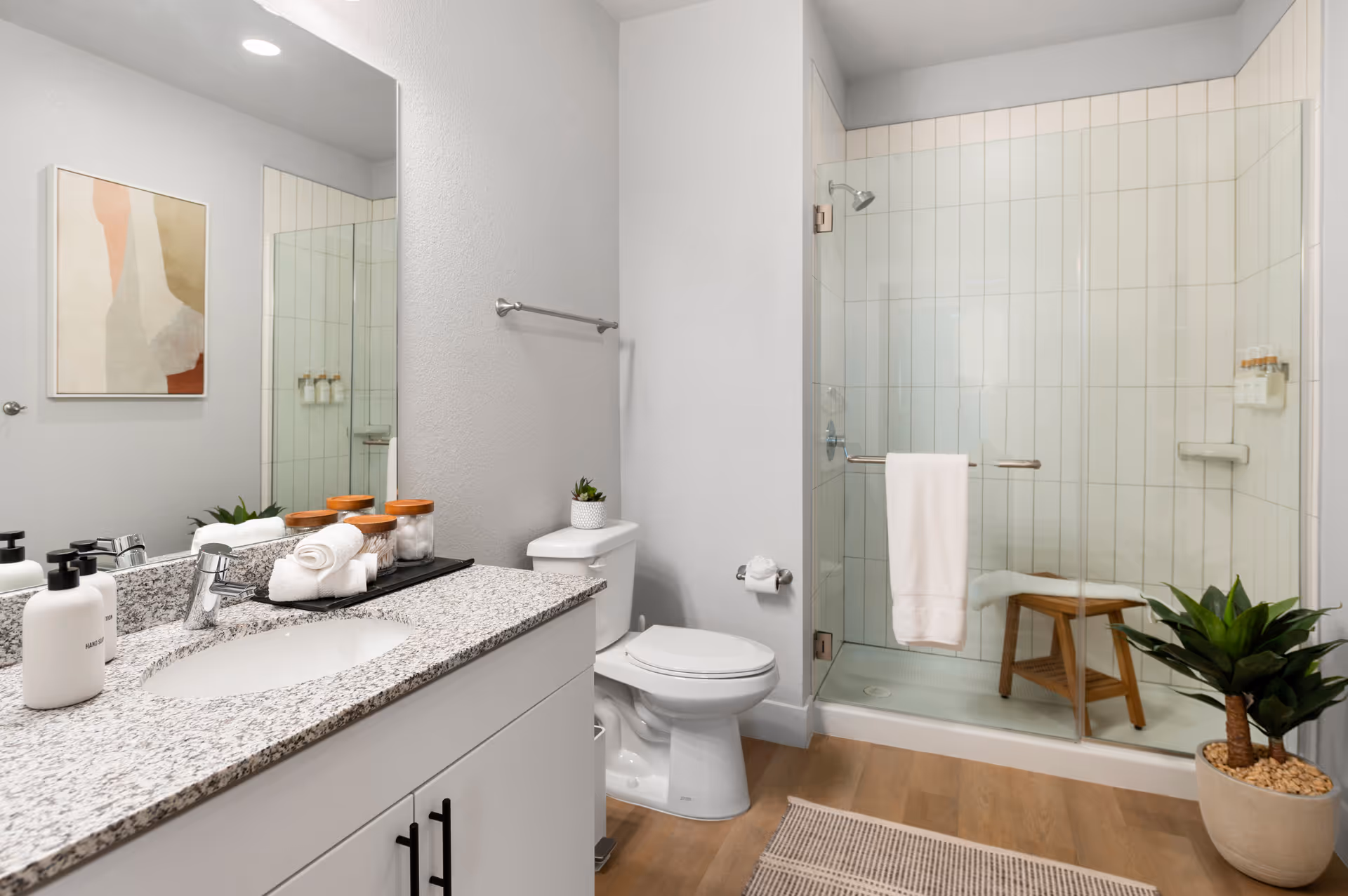 Bright modern bathroom with a granite-topped vanity, toilet, and a glass-enclosed shower with a wooden stool.