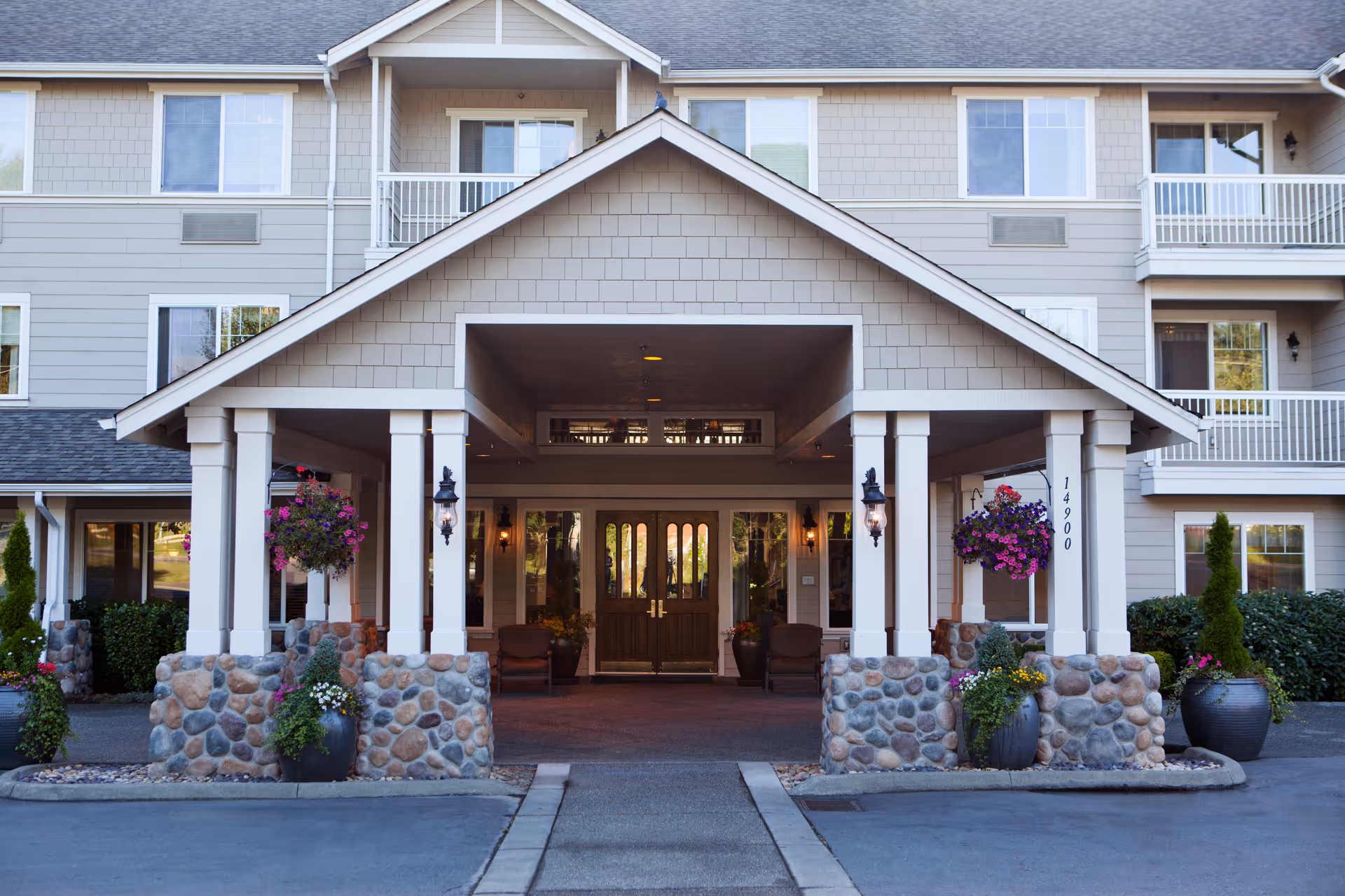 Front entrance of a senior living facility with a covered driveway supported by white pillars with stone bases. Hanging flower baskets and potted plants decorate the entrance area. The building has beige siding and multiple windows with balconies on the upper floors.