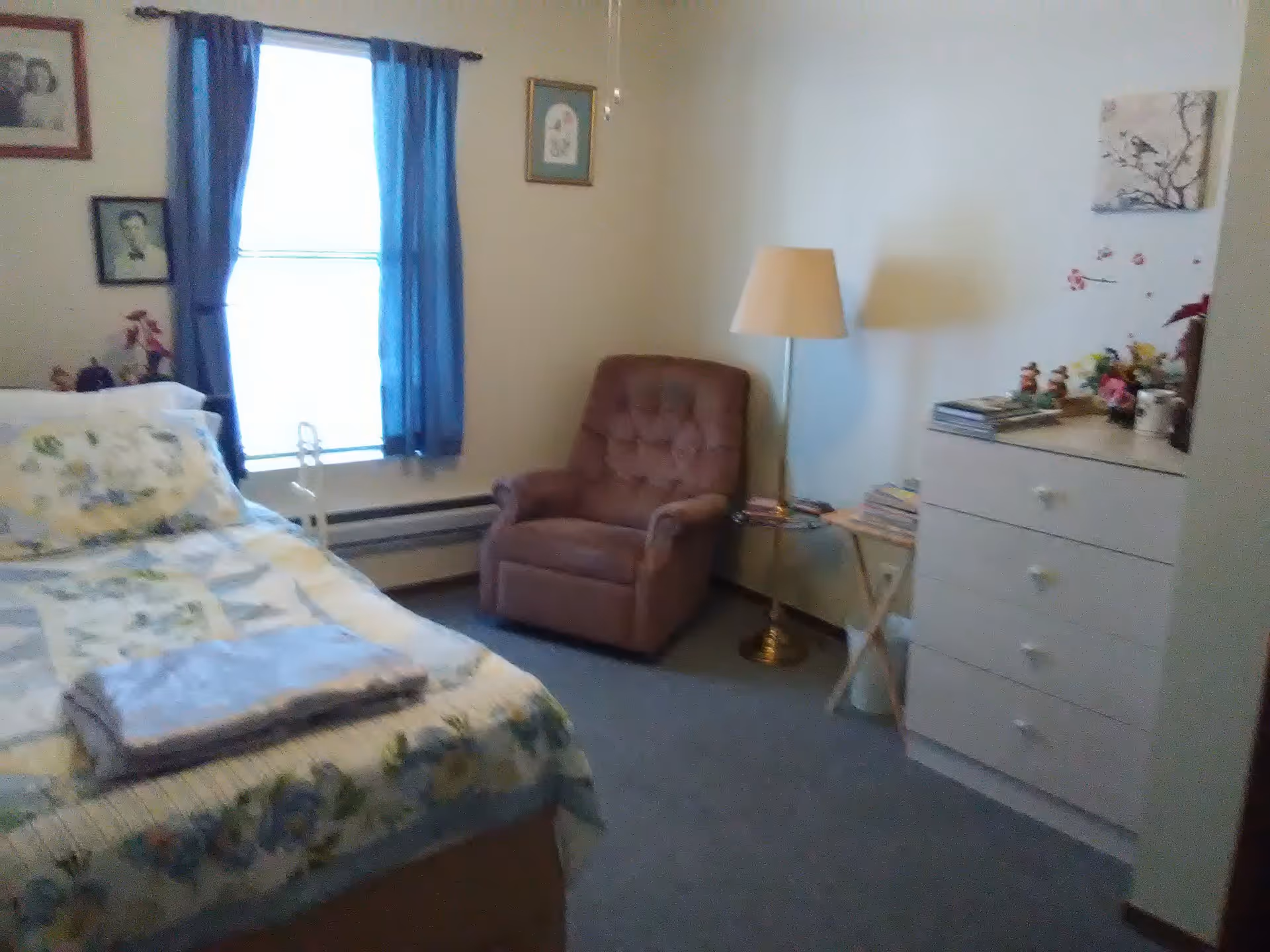 A cozy bedroom with a bed covered in a floral quilt and a folded towel on top. There is a brown upholstered armchair next to a window with blue curtains. A floor lamp stands beside a small wooden side table holding books. A white dresser with four drawers is against the wall, decorated with small figurines and flowers. The walls have framed pictures and a painting of birds on a branch.