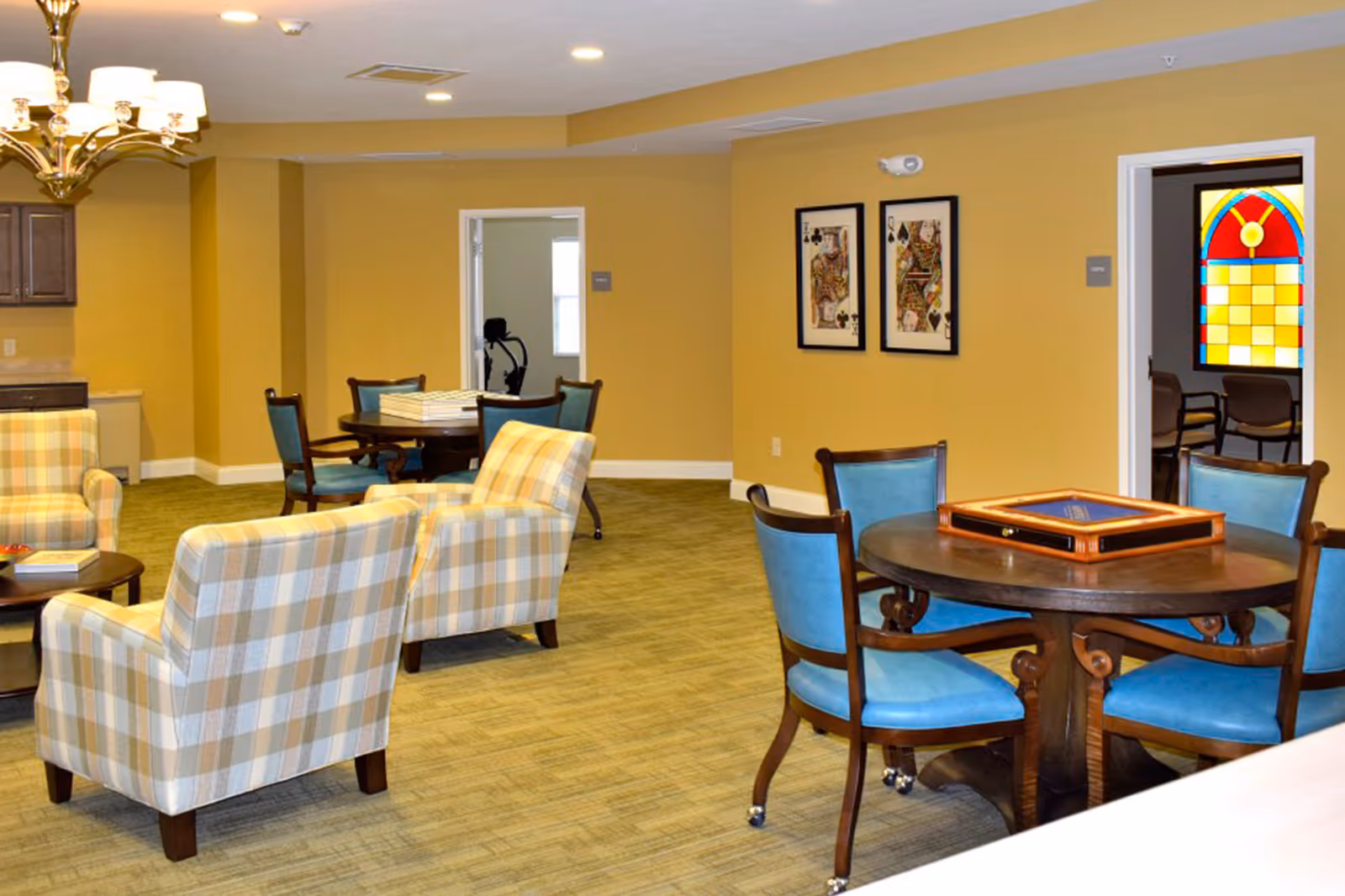 A cozy common area in a senior living facility with plaid upholstered armchairs, round tables with blue cushioned chairs, and framed playing card artwork on the wall. A stained glass window is visible through an open doorway, and a chandelier hangs from the ceiling.