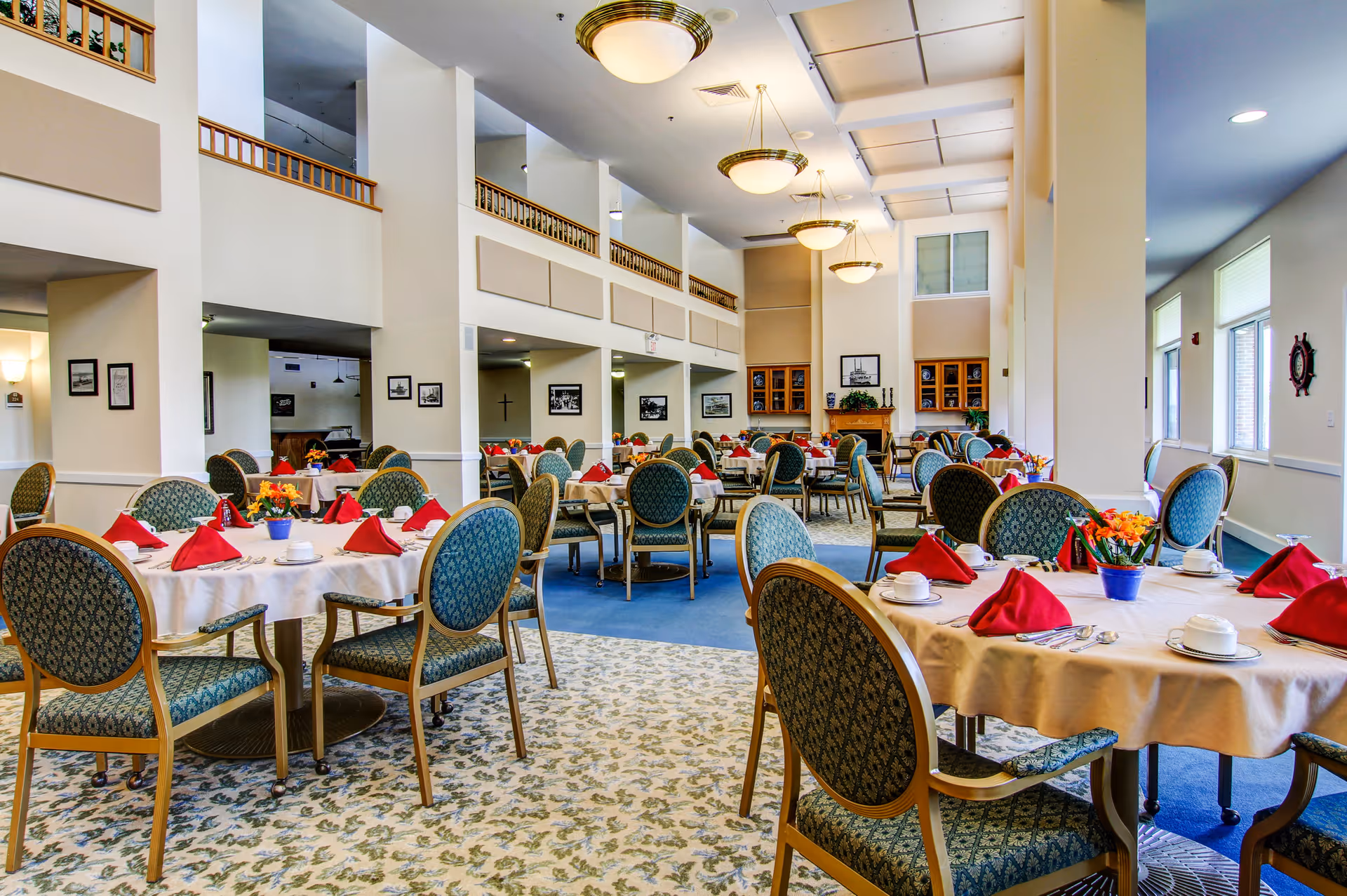 A spacious dining room in a senior living community with round tables covered in beige tablecloths, each set with white cups, saucers, silverware, and red folded napkins. The room features patterned upholstered chairs, large windows letting in natural light, and decorative ceiling lights. There are framed pictures on the walls and a fireplace with shelves in the background.
