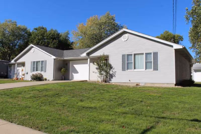 Single-story light-gray ranch-style building with an attached garage, green lawn, and trees under a clear blue sky.