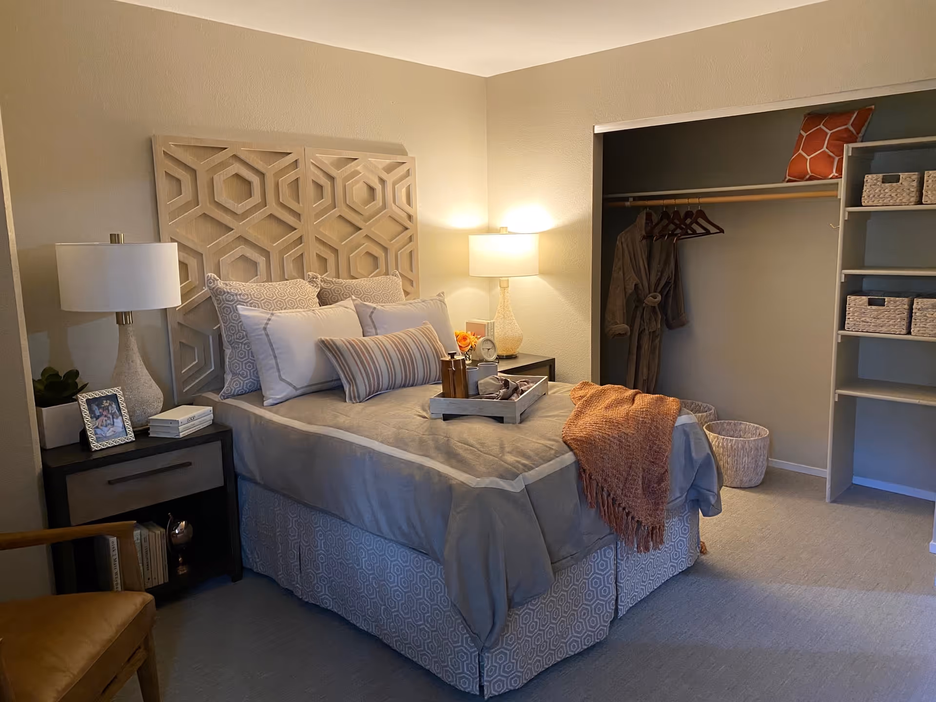 A cozy bedroom with a bed featuring a decorative wooden headboard, multiple pillows, and a tray with items on the bed. There are two bedside tables with lamps, a framed photo, books, and a plant. An open closet with a robe hanging and shelves with baskets is visible. A brown chair is partially visible in the corner.