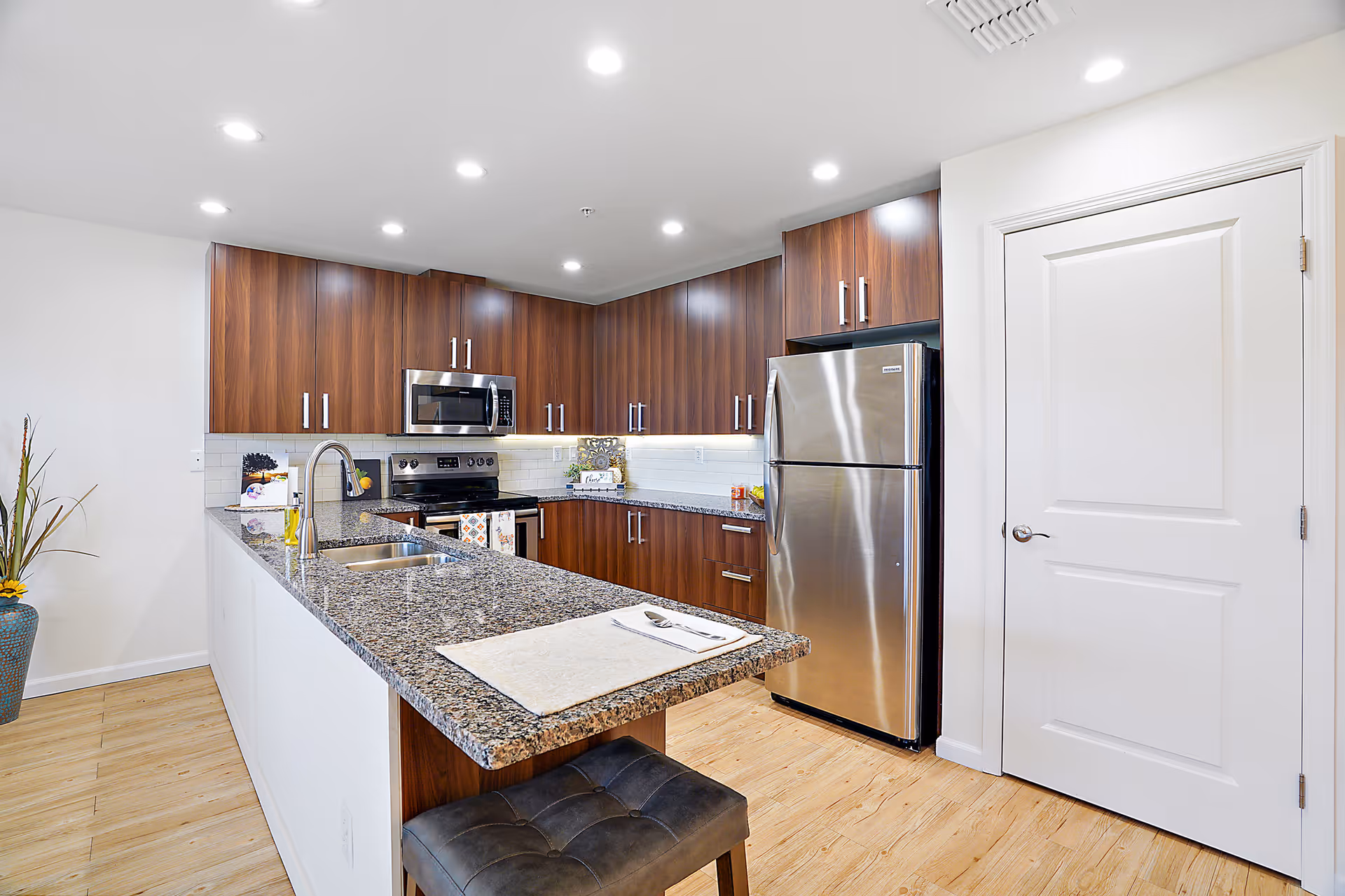 Modern kitchen with wooden cabinets, granite countertops, stainless steel refrigerator, oven, microwave, and a sink on the island. There is a stool at the island and a white door on the right side of the image.