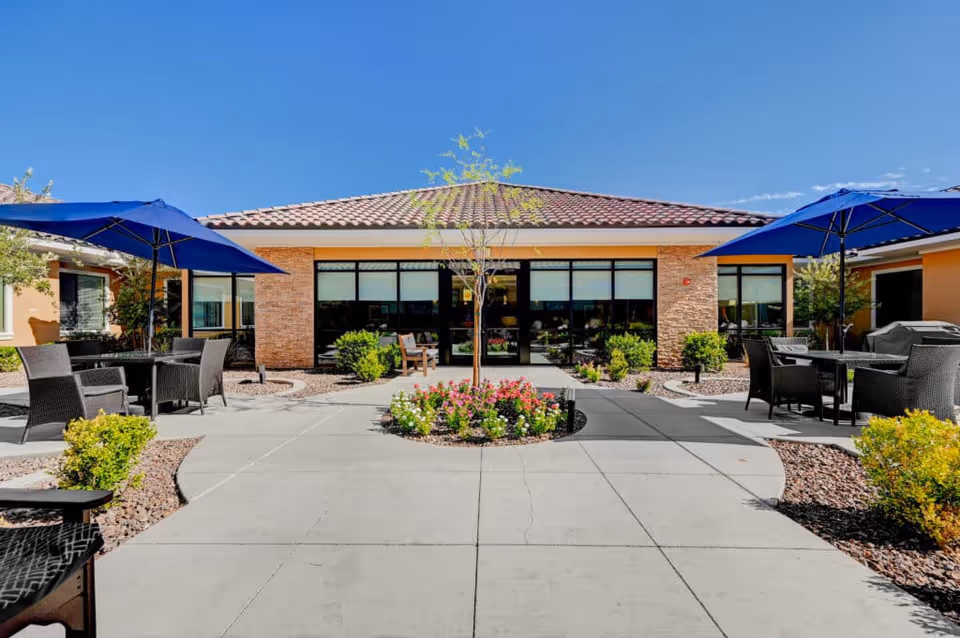 Sunny outdoor courtyard with patio tables and blue umbrellas, flower beds, and a single-story building entrance.