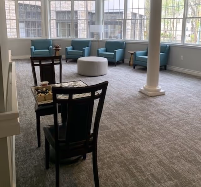 A spacious, carpeted common area with large windows letting in natural light. The room features four blue armchairs arranged in a semi-circle around a round white ottoman. In the foreground, there is a small table with two dark wooden chairs, and a decorative column stands near the center of the room.